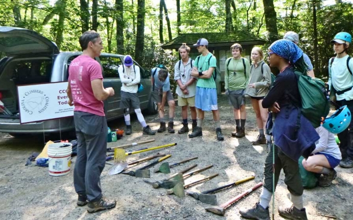A person speaks to a group of students. There are garden tools laying on the ground between them.